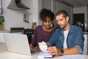 Young multiethnic couple checking bills while managing accounts on home banking app. Serious guy and african woman sitting at home discussing finance for the month. Young casual man and girl using laptop while looking at invoice and plan the budget to save.
